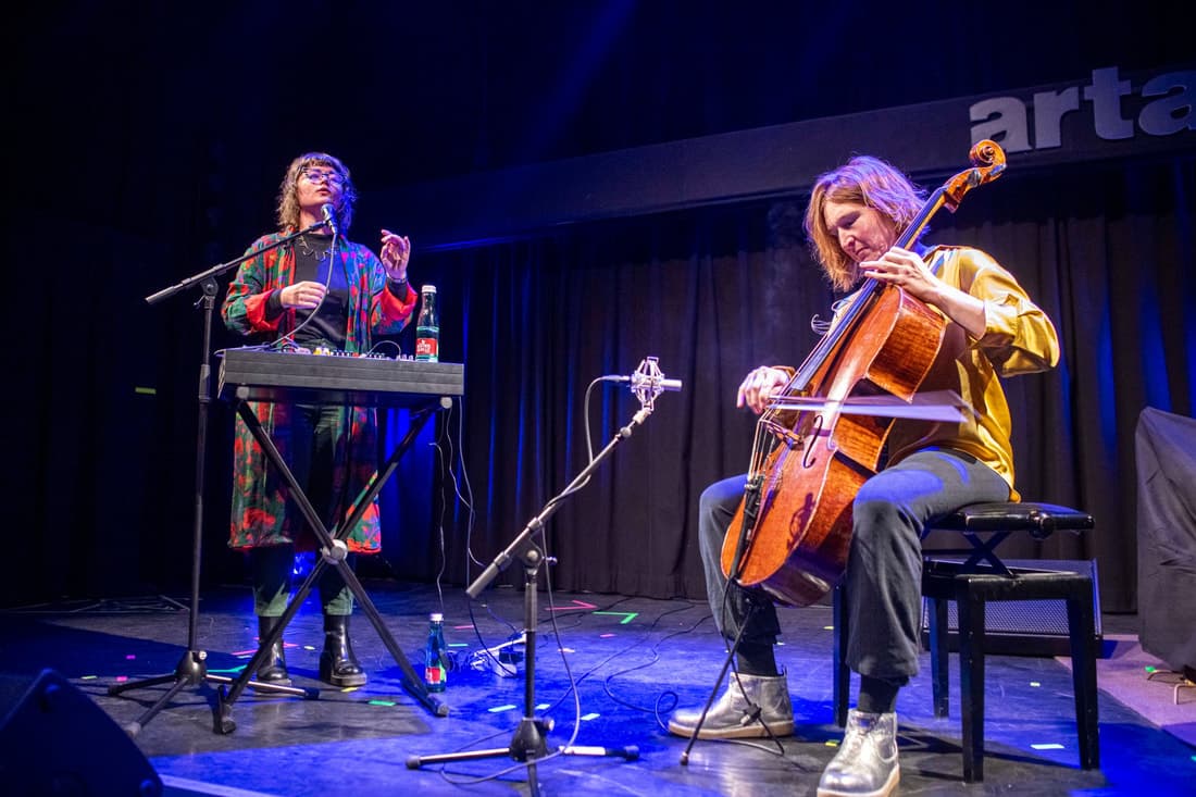 Two musicians perform on stage, one playing a cello and the other using a keyboard and microphone.